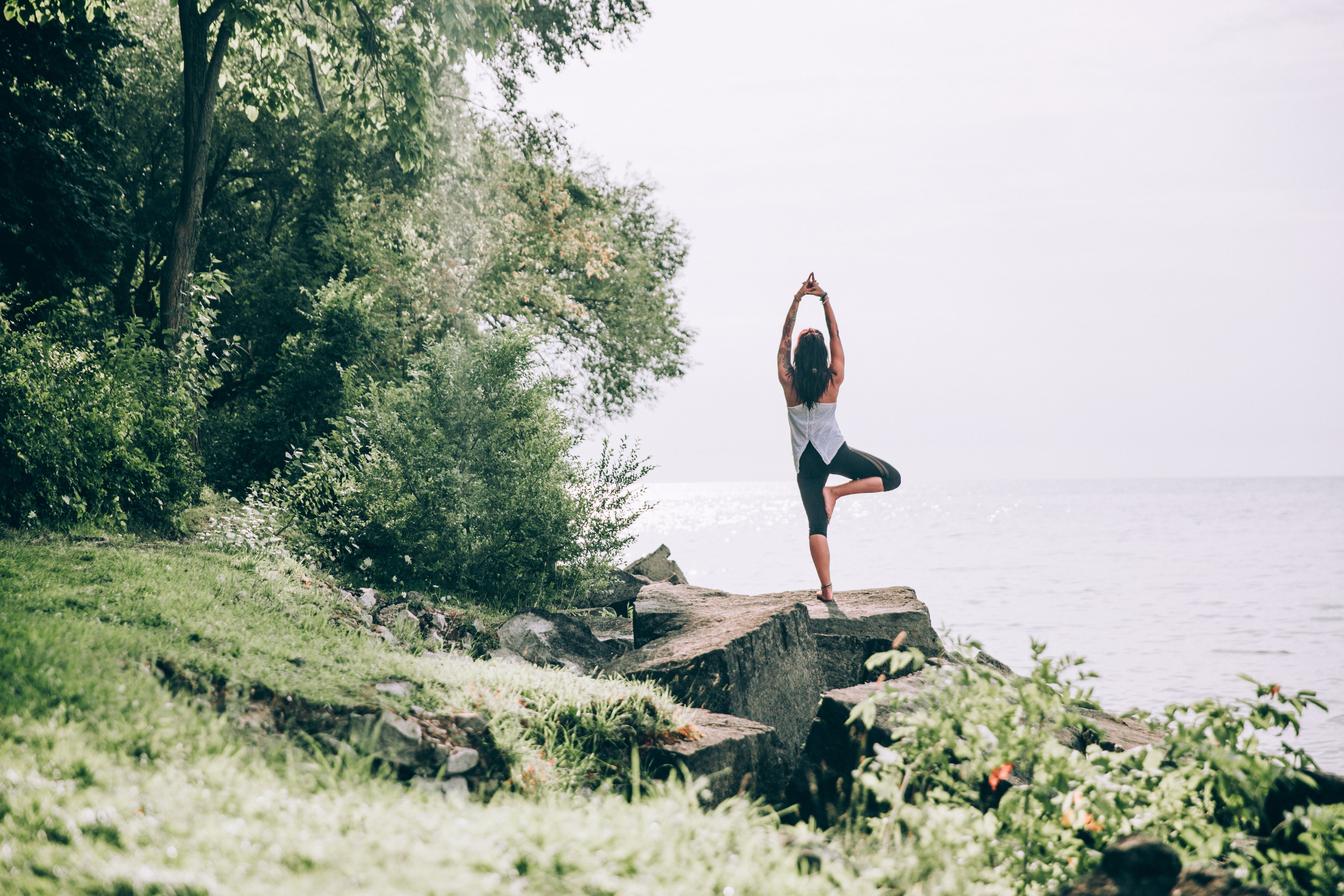 Woman standing on a rock ledge looking out over the ocean in a yoga pose with trees and forest to the left of her.  